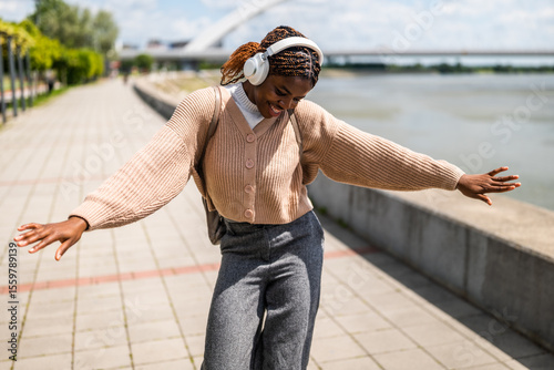 Happy young black woman is walking on foot path, listening music and smiling. She is enjoying sunny day outdoor.
