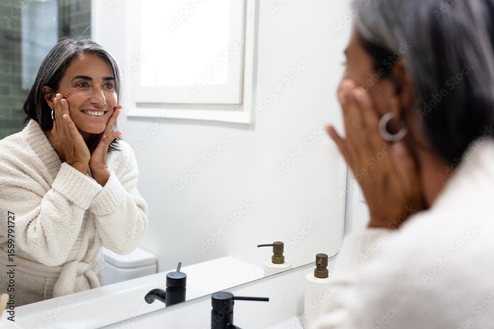 Mature happy woman touching and examining facial skin looking in bathroom mirror. Mature happy woman touching and examining facial skin looking in bathroom mirror.