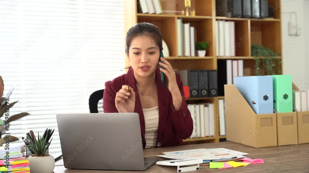 Focused Communication: An Asian professional, engrossed in a phone conversation while working at her desk, surrounded by the trappings of a modern office, exuding competence and dedication.