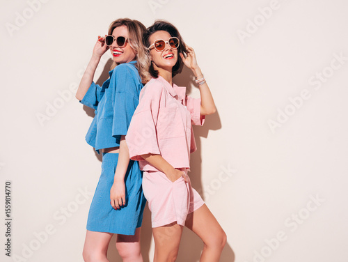 Two young beautiful smiling brunette and blond female in trendy summer clothes. Carefree women isolated, in studio. Positive models having fun. Cheerful and happy in sunglasses
