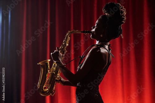 African American woman playing saxophone on stage in front of red curtain, standing in profile with hair in dreadlocks, performing jazz music as professional musician