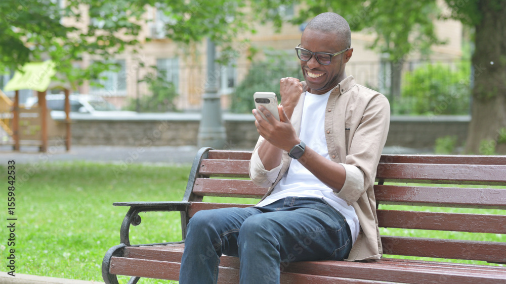 Obraz premium Young African Man Expressing Excitement While Using Phone on a Park Bench