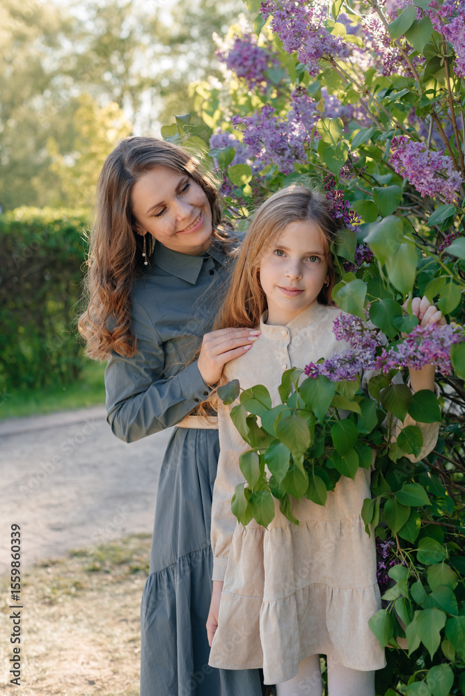 Fototapeta premium A happy mother and daughter in front of a blooming lilac tree. A family vacation on a sunny, warm day. Raising children, a happy family, and family values.