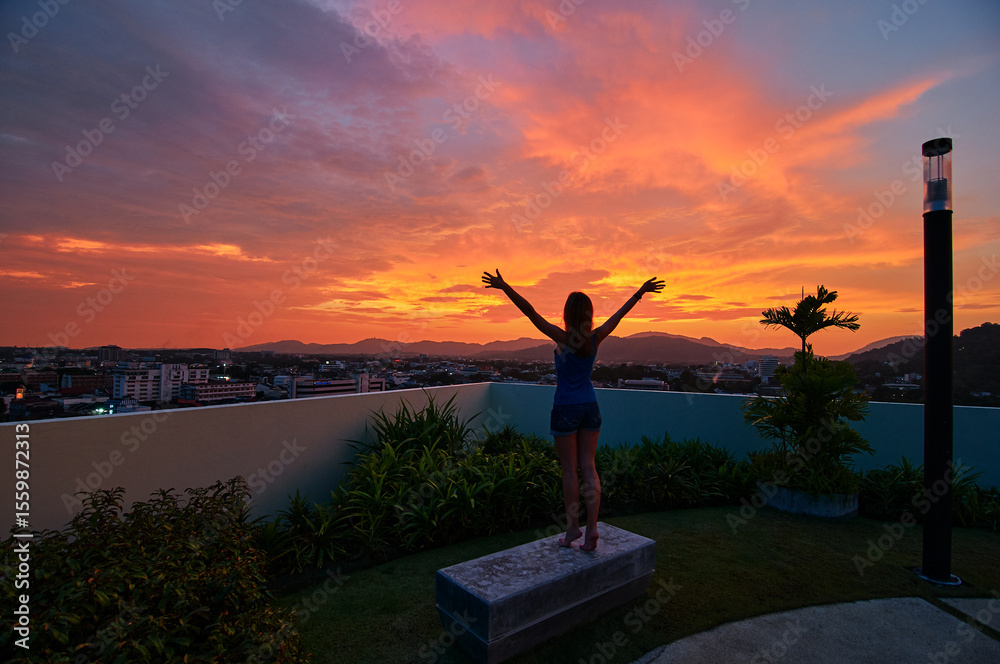 Fototapeta premium A person stands on a rooftop with arms raised, silhouetted against a vibrant sunset sky. The cityscape and distant mountains are visible in the background, creating a serene and uplifting scene.
