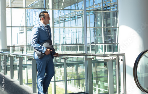 Businessman leaning on railing in atrium, holding jacket on forearm, overlooking escalator and lawn