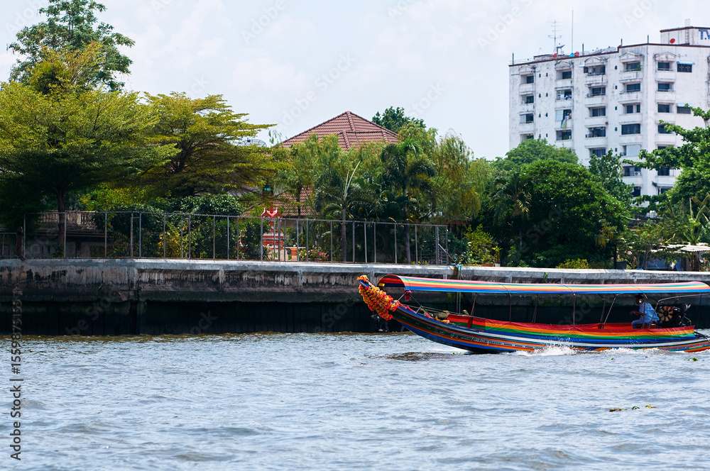 Obraz premium Colorful Longtail Boat Gliding by Riverside Urban Scenery Under Clear Sky