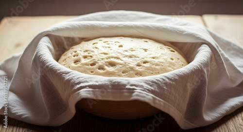 Bread dough rises in a bowl covered with a white cloth on a wooden table