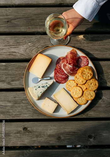 A rustic charcuterie board with assorted cheeses, crackers, and cured meats on a ceramic plate over a weathered wooden deck, with a hand holding a glass of white wine entering the frame. 