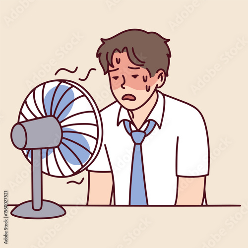Man sits at desk sweating and wipes forehead while fan blows. He struggles with heat and exhaustion in indoor workplace