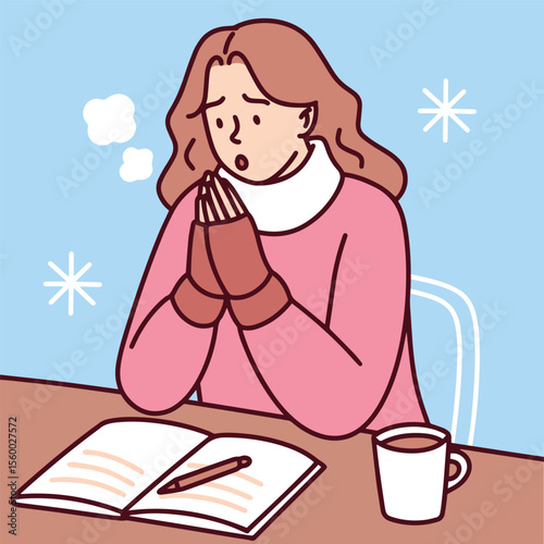 Young woman sits at table reading a book and warming hands near hot drink in cold unheated room