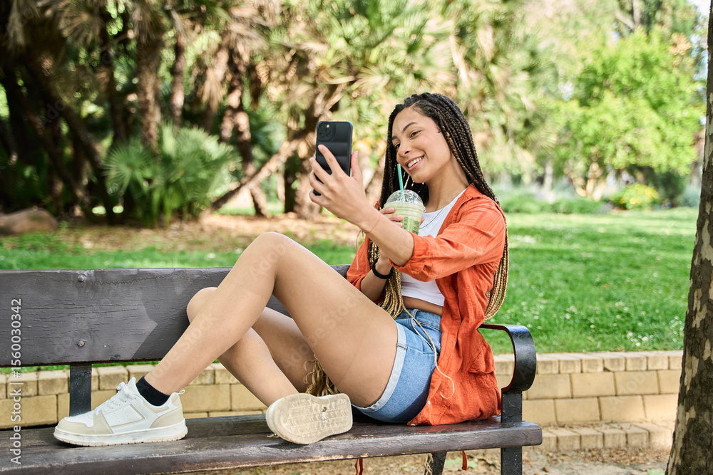 Naklejka premium Young latina woman taking a selfie while enjoying a refreshing drink in the park
