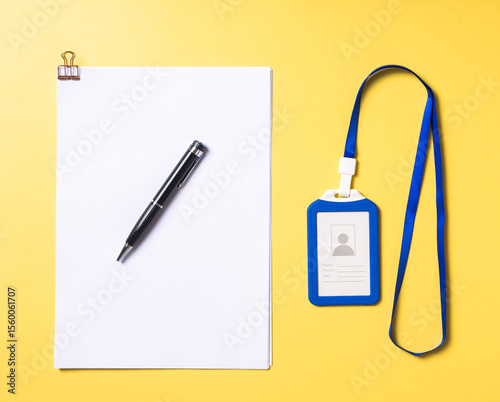 Top view of a blue ID card with lanyard, a black pen on top of clipped white papers, all neatly arranged on a bright yellow background