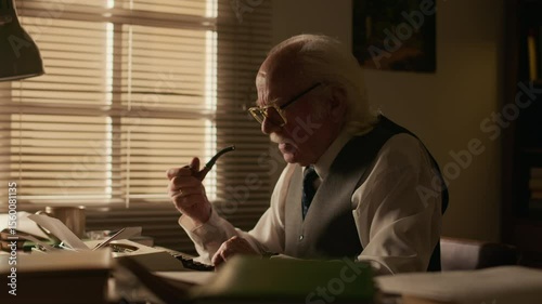 Side view medium shot of concentrated senior Caucasian male novelist writing new chapter using vintage typewriter while smoking pipe sitting at desk in his retro study room