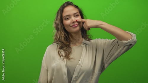 Hispanic young woman with long curly hair points finger to nose in studio with green screen; odor and disgust.