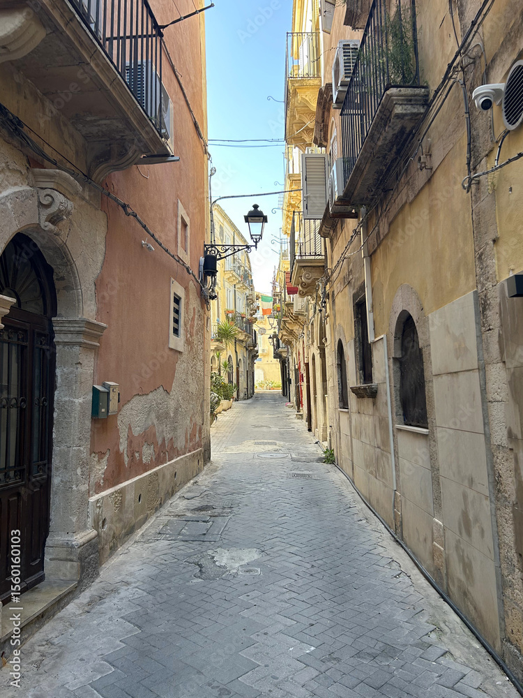 Obraz premium A narrow street lined with old stone buildings and balconies winds through a quiet European town. Potted plants, shuttered windows, and warm sunlight create a charming, historic atmosphere.
