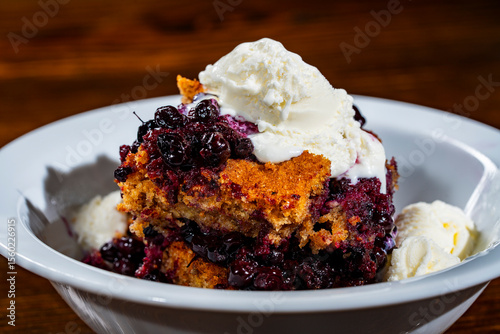 White plate with piece of fresh baked blueberry cobbler and ice cream on wooden background, closeup. Homemade berry cobbler ready to eat