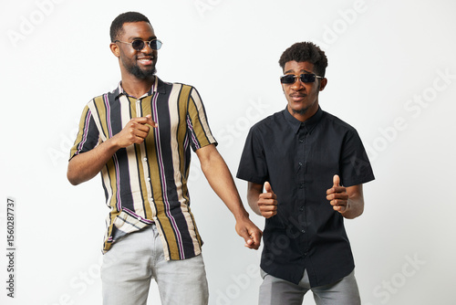 Two cheerful Black men dance joyfully together, wearing stylish sunglasses and colorful shirts, against a bright white background, showcasing friendship and celebration.