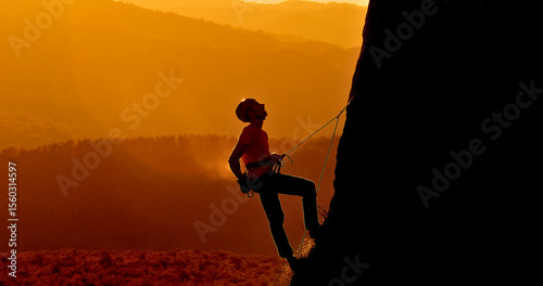 A climber in a red shirt and helmet scales a steep rock face at sunset. The silhouette of the climber contrasts against the warm, golden sky.