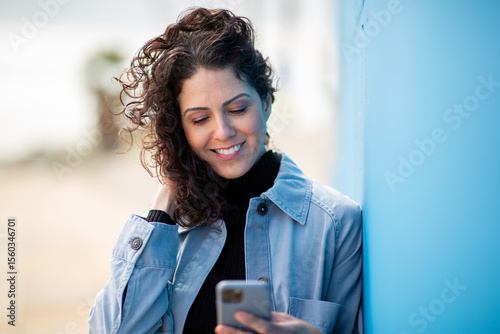 Papier peint Happy woman using smartphone, leaning against blue wall with windblown hair