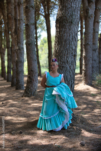 A beautiful young Spanish woman dressed in a traditional green flamenco dress with a flower in her hair rests leaning against a pine tree at a typical pilgrimage in Andalusia the woman holds her dress photo