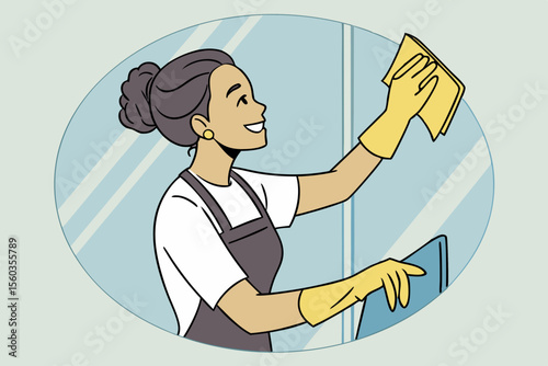 Smiling female cleaner wearing gloves wiping glass window with yellow cloth
