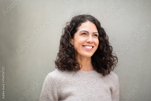 Portrait of a cheerful woman with curly hair standing against a light gray wall, smiling brightly and looking away