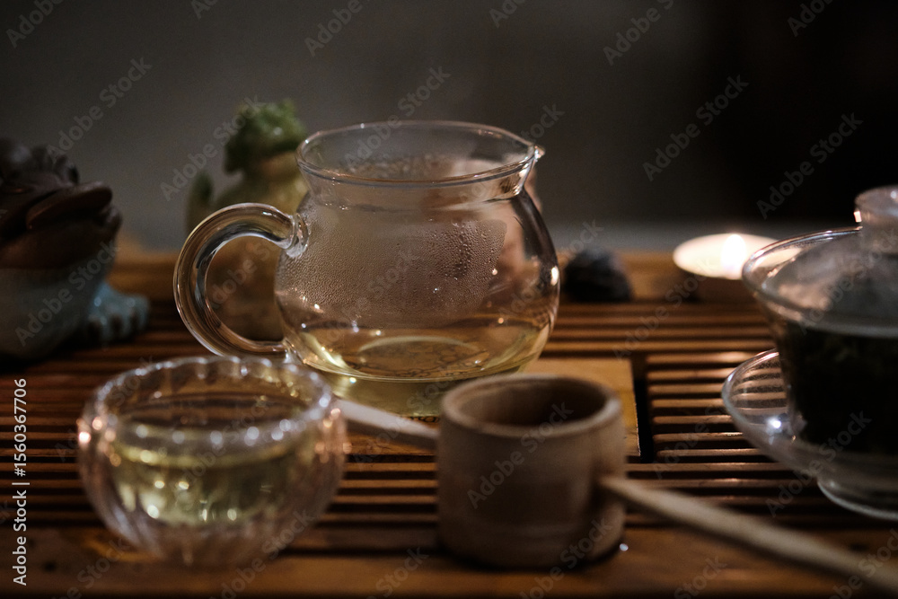 Fototapeta premium Warm and intimate scene of traditional tea ceremony arrangement on wooden tea tray. Transparent glass teapot with warm tea, matching cup, bamboo strainer and gaiwan with tea leaves placed in harmony