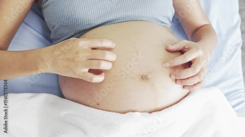 Pregnant Asian woman sits on hospital bed gently scratching and rubbing her belly with both hands relieve itching caused by skin expansion dry cracking and rashes waits doctor to check her pregnancy.
