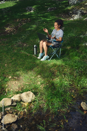 Woman Relaxing Outdoors Using a Laptop in a Natural Setting by a Calm Stream
