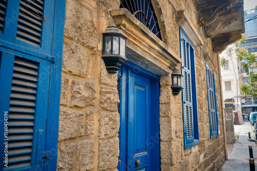 Traditional Mediterranean-style stone building with vibrant blue wooden shutters and a matching door. Black metal lanterns frame the entrance