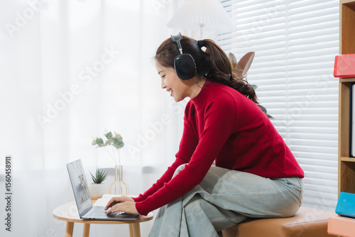Focused Harmony: An Asian woman is deeply engrossed in her task, seated by a window, radiating concentration and calmness while enjoying the immersive sound of headphones and engaging with technology.