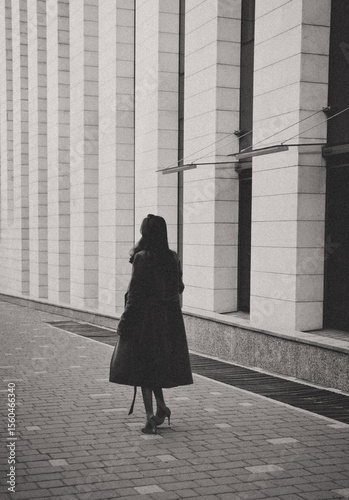 A woman in a long coat and high heels walks alone by a modern building with tall columns, captured in a black and white photo