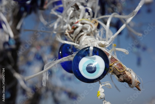 Traditional Turkish evil eye glass charms (nazar boncuğu) tied to tree branches with white string against a bright blue sky. Believed to ward off bad luck and attract protection.