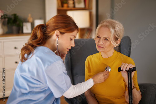 A home health aide in light blue scrubs listens to an elderly woman's heart with a stethoscope. The senior woman sits comfortably with a walking cane in her hand in her home.