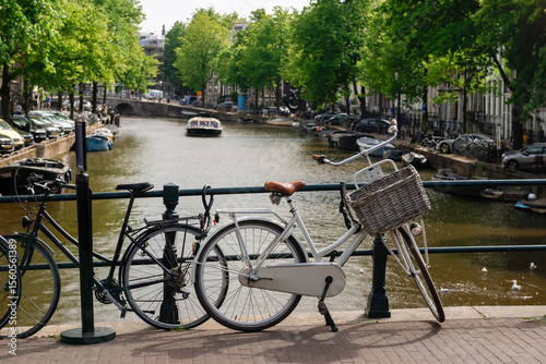 Two bicycles, one with a wicker basket, parked on a bridge overlooking an Amsterdam canal with boats and trees