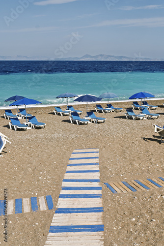 Fototapeta Naklejka Na Ścianę i Meble -  sand beach with blue deck chairs and umbrellas arranged in rows. A wooden walkway leads from the foreground to the sea turquoise in Rhodes, Greece