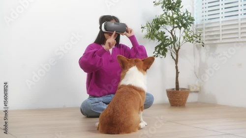 A young Asian woman relaxes indoors in an empty room, wearing VR glasses while her playful Corgi dog stays beside her. The moment captured companionship, technology, and the joy of togetherness.
