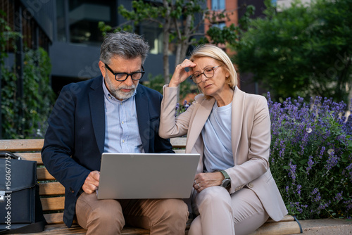 Valokuvatapetti Worried businessman and businesswoman reviewing bad news and poor project results on laptop while sitting outdoors in urban business district