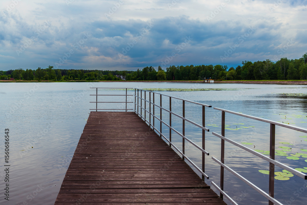 Obraz premium A scenic view of a wooden pier with metal railings extending into a tranquil lake surrounded by lush green trees. Lily pads float gently on the surface, and a moody, cloudy sky adds atmosphere to the 