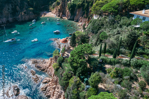 An aerial view showcases the stunning Marimurtra Botanical Garden, with its diverse flora, charming gazebo, and elegant villa overlooking the clear blue Mediterranean Sea.