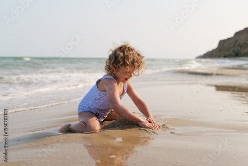 A cheerful young girl with curly hair in a swimsuit plays actively with sand and water at the beach.