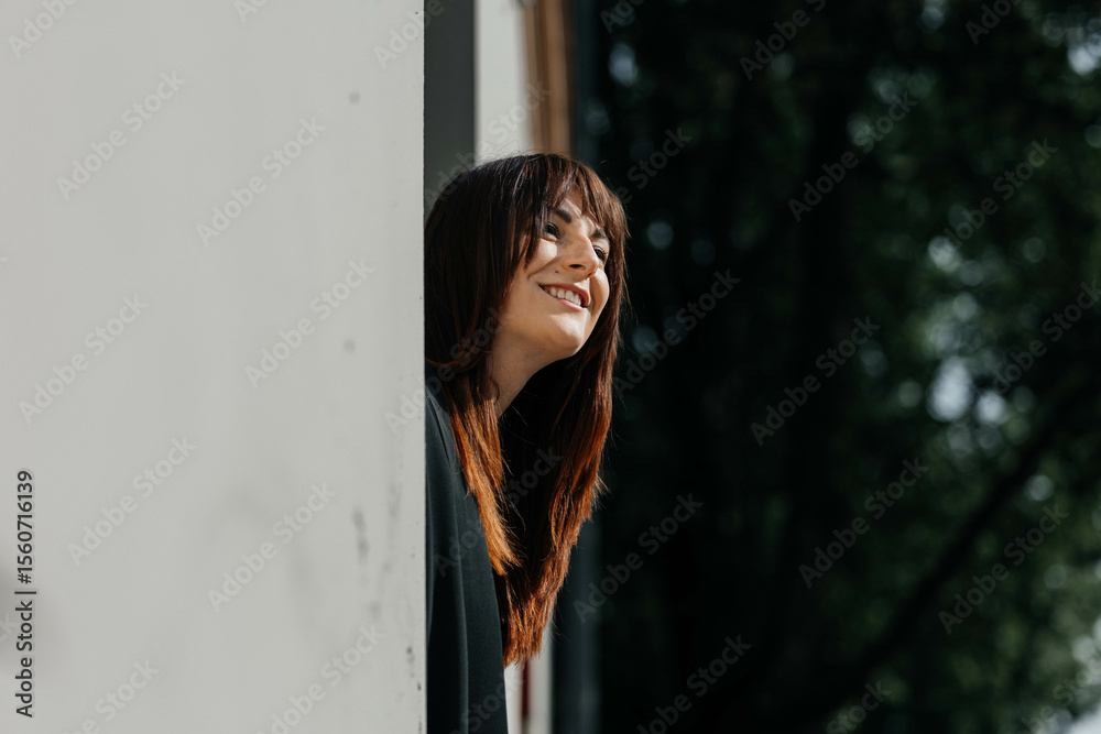 Fototapeta premium Young caucasian female smiling while leaning out a window in natural light