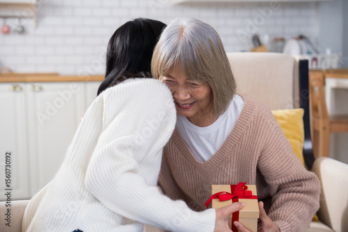 Daughter giving a gift to her elderly mother, who smiles happily and hugs her daughter on the living room sofa, showing love, family bonding, and warmth in a cozy home setting.