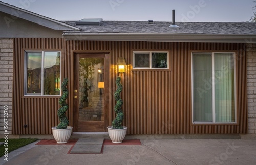 Exterior of a mid-century house with wood paneling, large windows, a porch light and spiral-shaped trees framing the entrance.