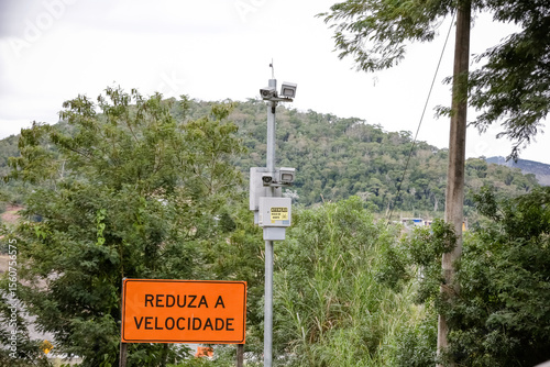 Radar fixo com placa “Reduza a Velocidade” em estrada cercada por vegetação. Imagem sobre segurança no trânsito e controle de velocidade em áreas rurais.