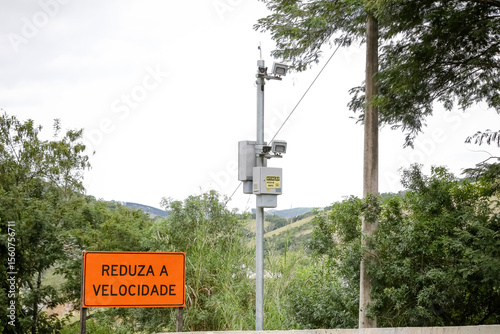 Radar fixo com placa “Reduza a Velocidade” em estrada cercada por vegetação. Imagem sobre segurança no trânsito e controle de velocidade em áreas rurais.