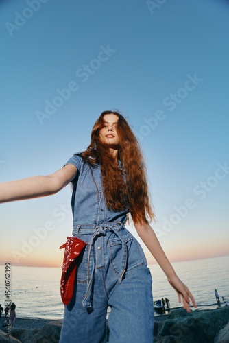 Lifestyle woman in modern boho-western denim street style with long hair by the sea during golden hour, film color enhancing casual youthful vibe and relaxed pose.