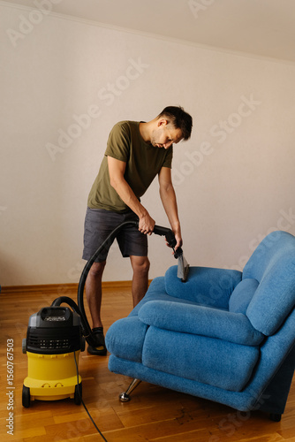 Full length vertical portrait of man in causal clothing using washing vacuum cleaner to clean blue sofa in home setting, demonstrating modern approach to household chores and upholstery maintenance.