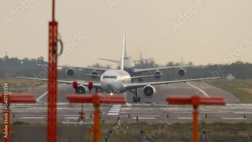 Two unmarked commercial airplanes taxi one behind the other on the runway, captured from a front view. The aircraft move slowly in line, preparing for takeoff. Ideal for themes of air travel