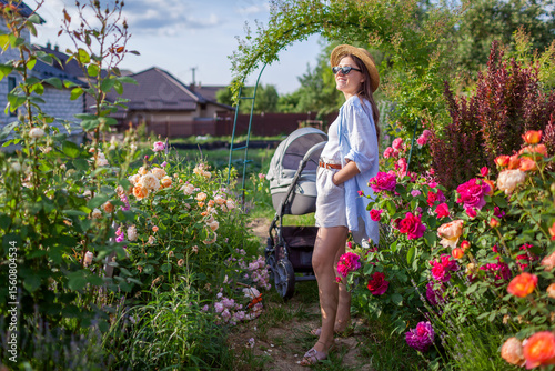 Happy young mother walking in her garden with stroller enjoying flowers. Woman gardener doing gardening chores while baby sleeps